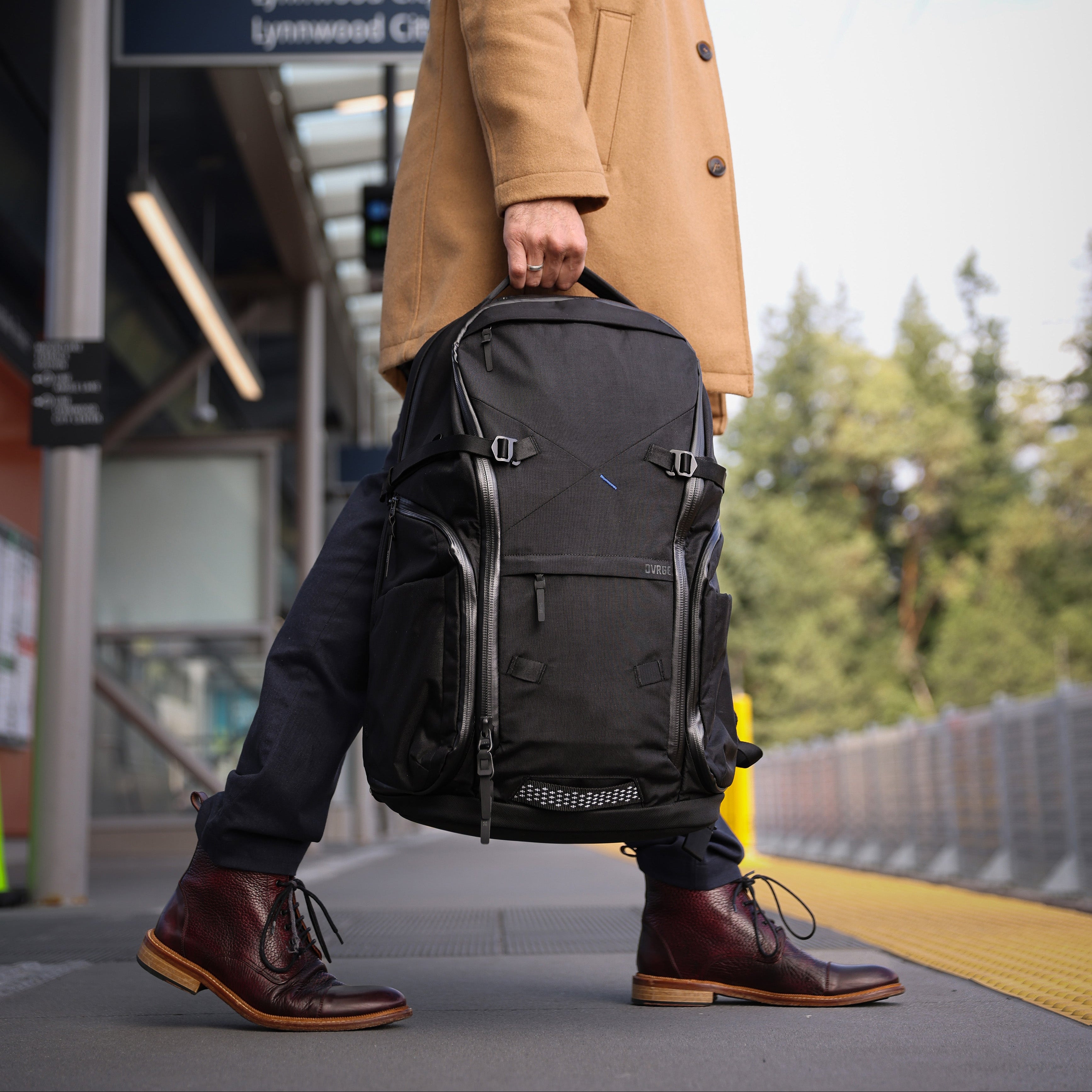 Man in a tan coat pulling a black backpack at a train station.